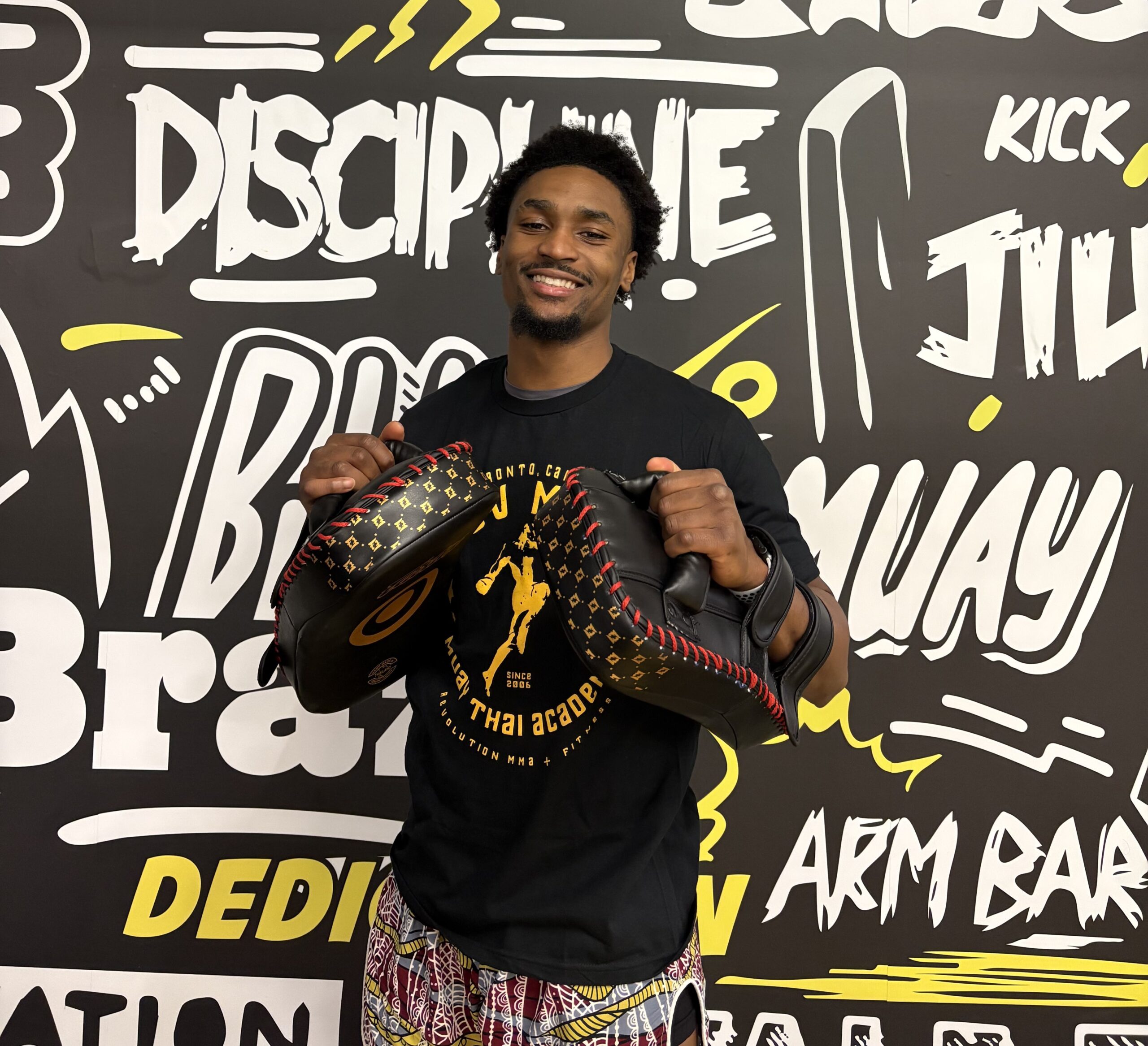 Smiling man holding boxing pads in front of a graffiti wall, promoting a Muay Thai academy.