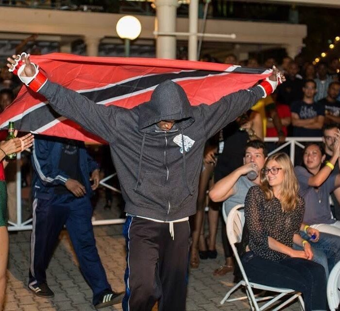 Boxer enters the arena with Trinidad and Tobago flag, crowd applauds enthusiastically in casual setting.