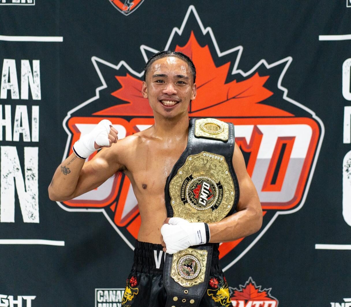 Muay Thai champion celebrates with title belt at Canadian Muay Thai Open, smiling victoriously in front of event banner.