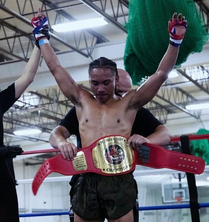 Boxer celebrates victory in ring, wearing champion belt, arms raised triumphantly, referee holding his hand.