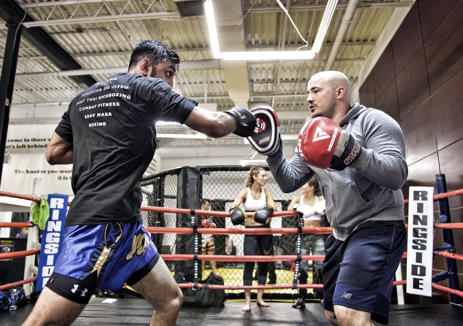 Two men sparring in a boxing ring at a gym, with onlookers in the background.