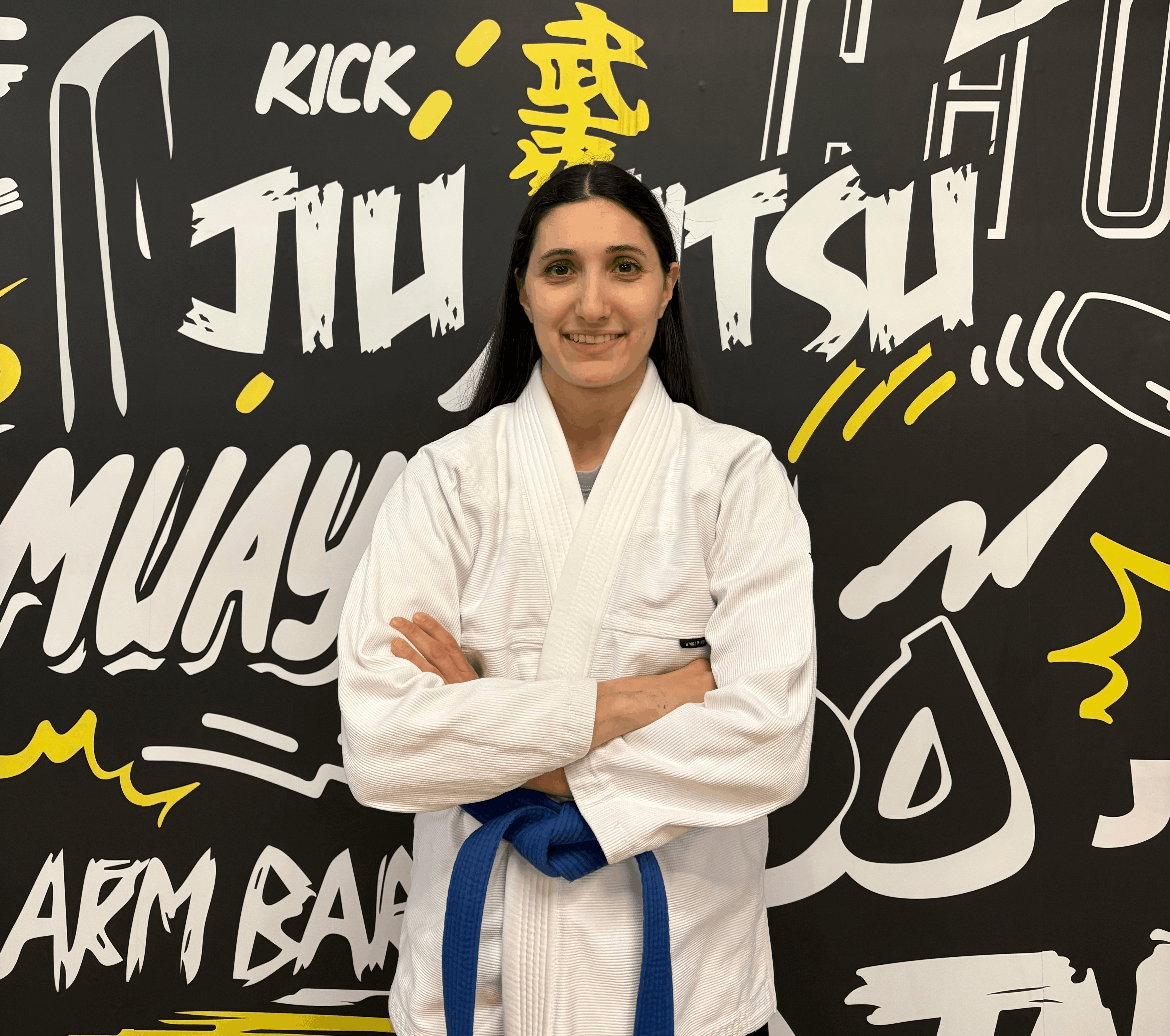 Person in a martial arts gi with a blue belt, standing in front of a graffiti wall with jiu-jitsu text.