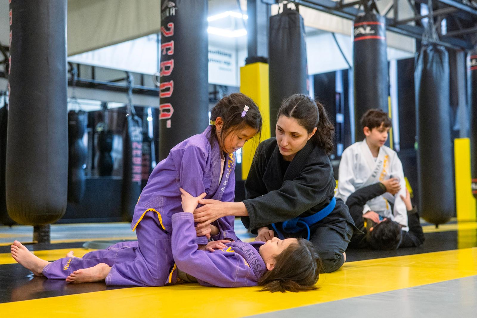Child practicing Brazilian jiu-jitsu with instructor in a gym, surrounded by punching bags and other students.