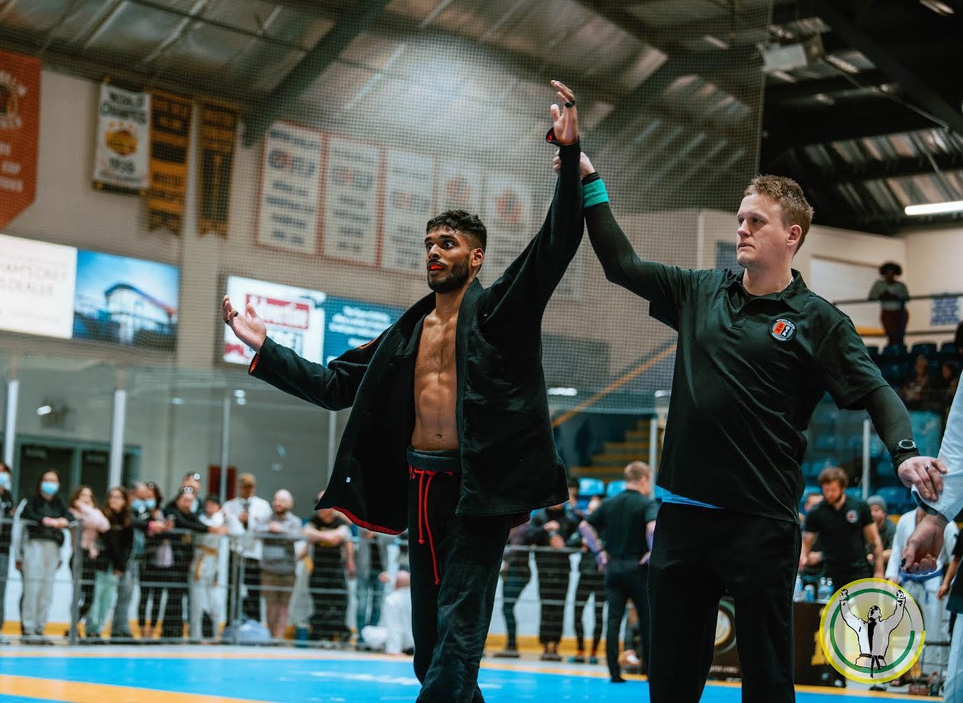 Martial arts competitor wins match, referee raises arm in victory, indoor arena with audience in background.