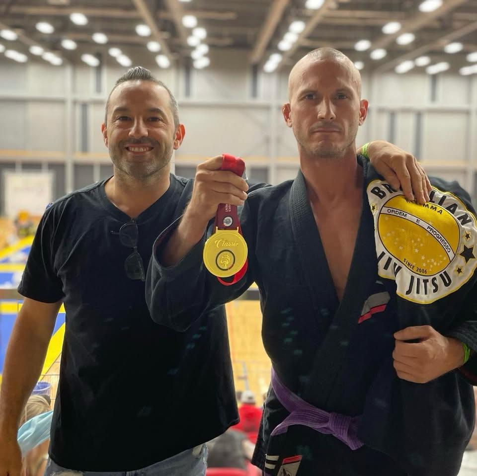 best-rated-bjj-classes-toronto-revmma Two men at a Brazilian Jiu-Jitsu event, one holding a gold medal, smiling and wearing martial arts attire.