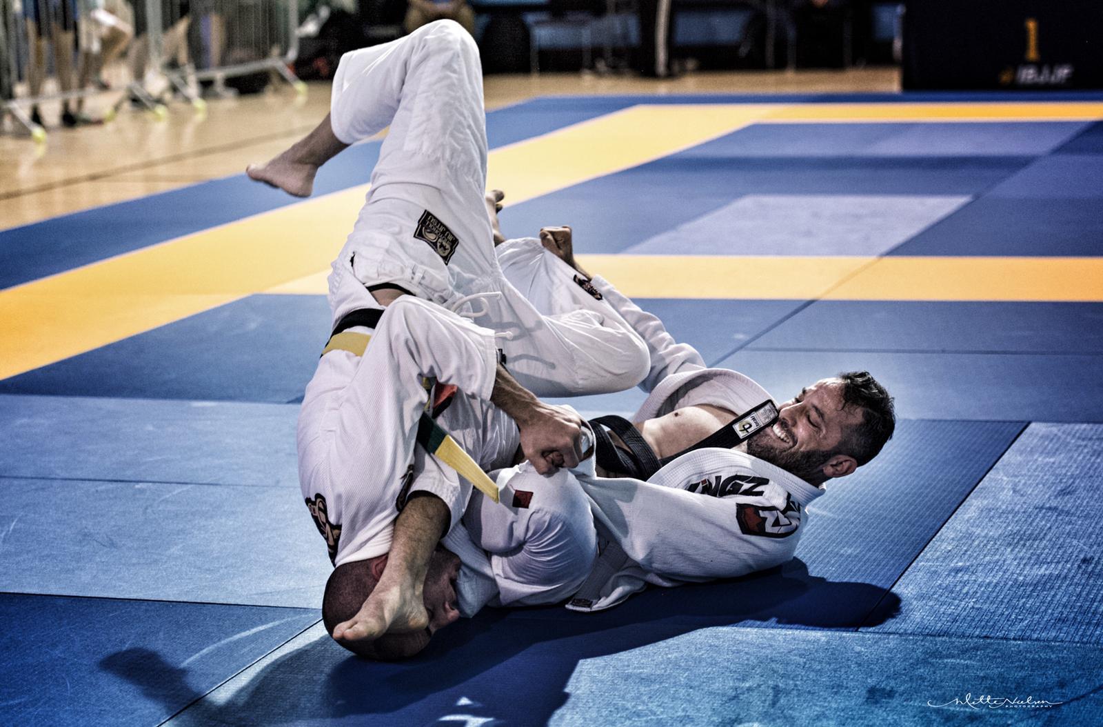 brazilian-jiu-jitsu-revmma Brazilian Jiu-Jitsu competitors in white gis grappling on a blue mat at a tournament.