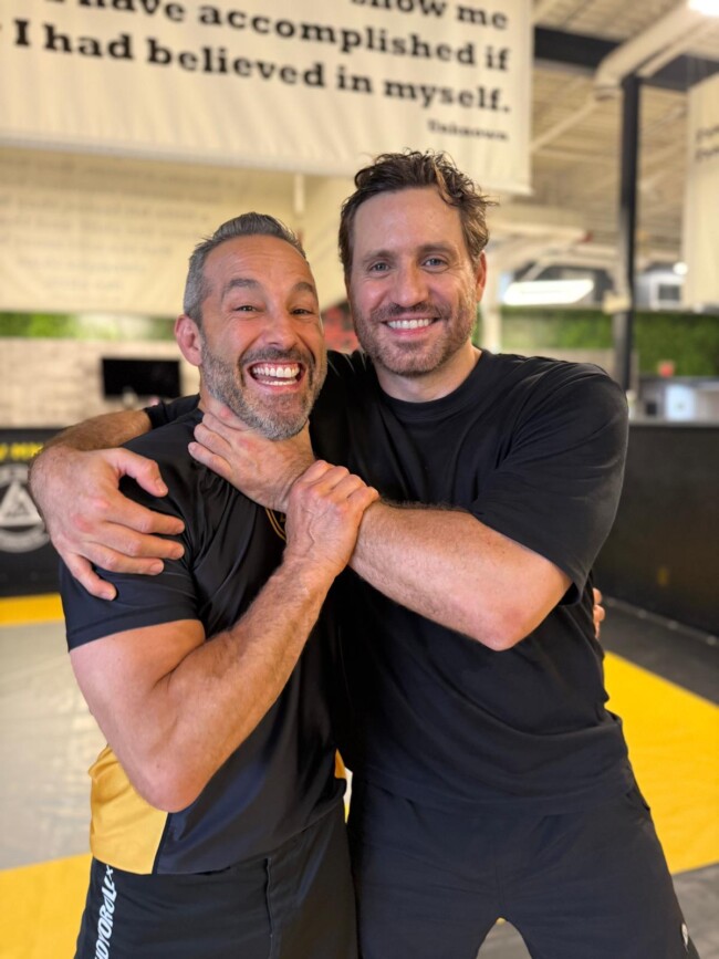 Joel training Hollywood actor Edgar Ramirez in Jiu Jitsu at RevMMA for his role in a Disney pilot. Two men smiling and posing in a gym, wearing sportswear, with motivational quote in the background.