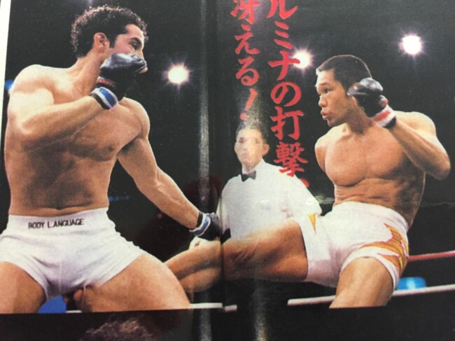 Joel fighting Rumina Sato, Japan 1998. Two martial artists in a kickboxing ring exchanging kicks, with a referee observing the action in the background.