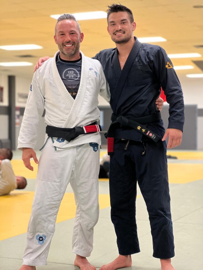 Joel training with World Champion Isaac Doederline. Two men in martial arts gis with black belts, posing in a dojo with an open mat setting.