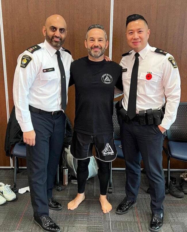 Joel with Chief of Police brass while training York Region officers at York Region Headquarters. Three individuals smiling, two in police uniforms, one in martial arts attire, standing in a room.