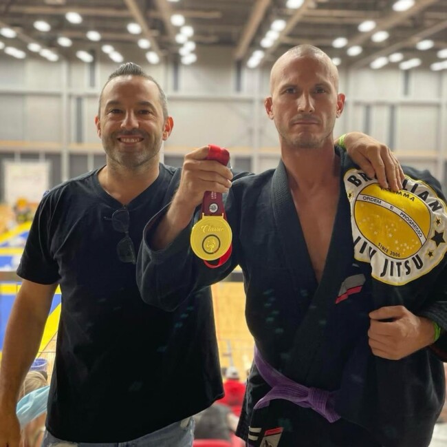 Joel with one of his personal students after winning Gold. Two men at a jiu-jitsu event, one holding a gold medal and wearing a black gi and a Brazilian jiu-jitsu patch.
