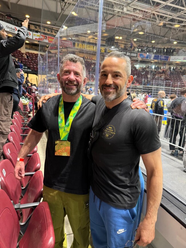 Joel with his personal student after winning the Ontario Open. Two smiling men pose together at a sports event; one wears a medal and the other a black t-shirt with Jiu-Jitsu.