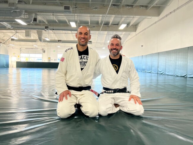 Joel training with Ryron Gracie, Gracie University, Los Angeles. Two men in Brazilian Jiu-Jitsu uniforms smiling and kneeling on a martial arts mat in a spacious gym.