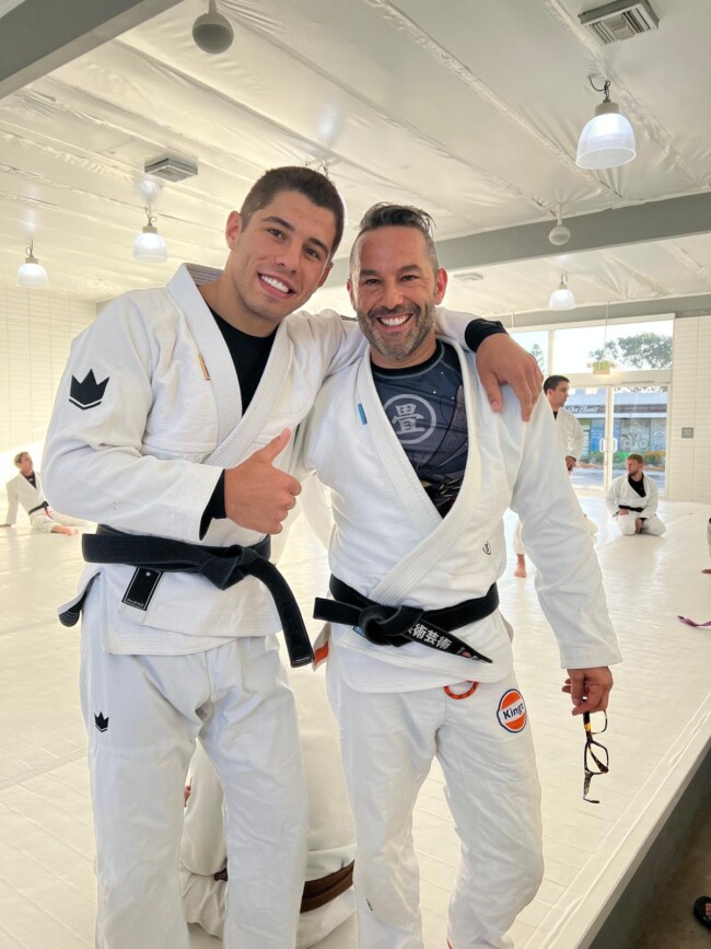 Joel training with The best jiu Jitsu athlete in the world and multiple time World Champion, Tainan Dalpra at Art of Jiu Jitsu, California. Two men in martial arts gis smiling and posing in a dojo, surrounded by others training in the background.