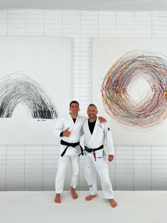 Joel training with 4X World Champion Gui Mendes, at Art of Jiu Jitsu Academy, California. Two martial artists in white gis with black belts, smiling in front of abstract art on a white brick wall.