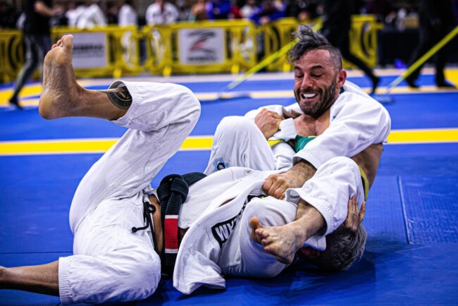 Joel winning via arm bar submission, World Masters Championship. Two martial artists in a Brazilian Jiu-Jitsu match on a blue mat, both wearing white gis, competing intensely.