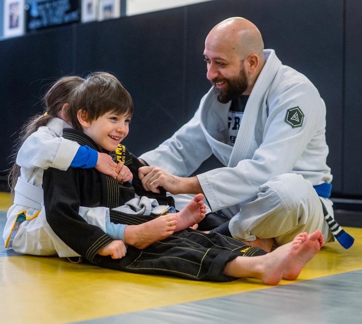 Kids practicing Brazilian Jiu-Jitsu with an instructor on the mat, smiling and learning grappling techniques.