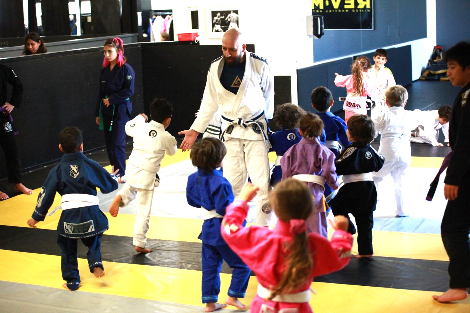 danial-bjj-kids-coach-revmma Children practicing Brazilian Jiu-Jitsu with an instructor on a dojo mat.