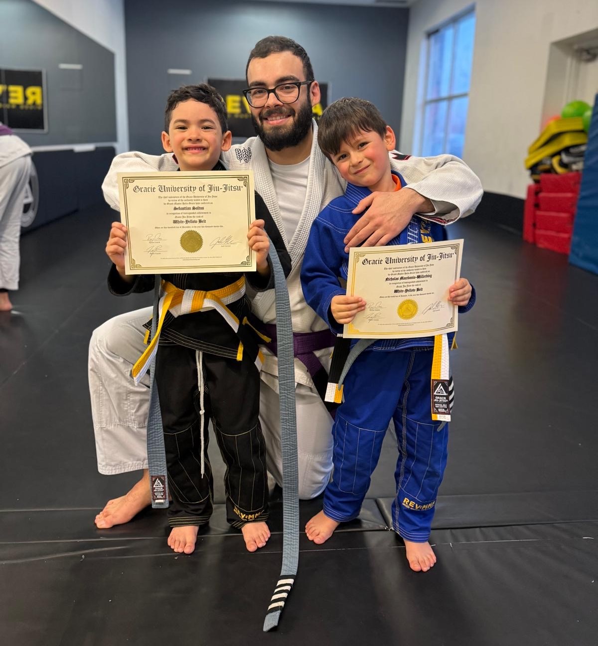 jiu-jitsu-philosophy-revmma Two children with certificates and an adult in martial arts uniforms at a jiu-jitsu academy.