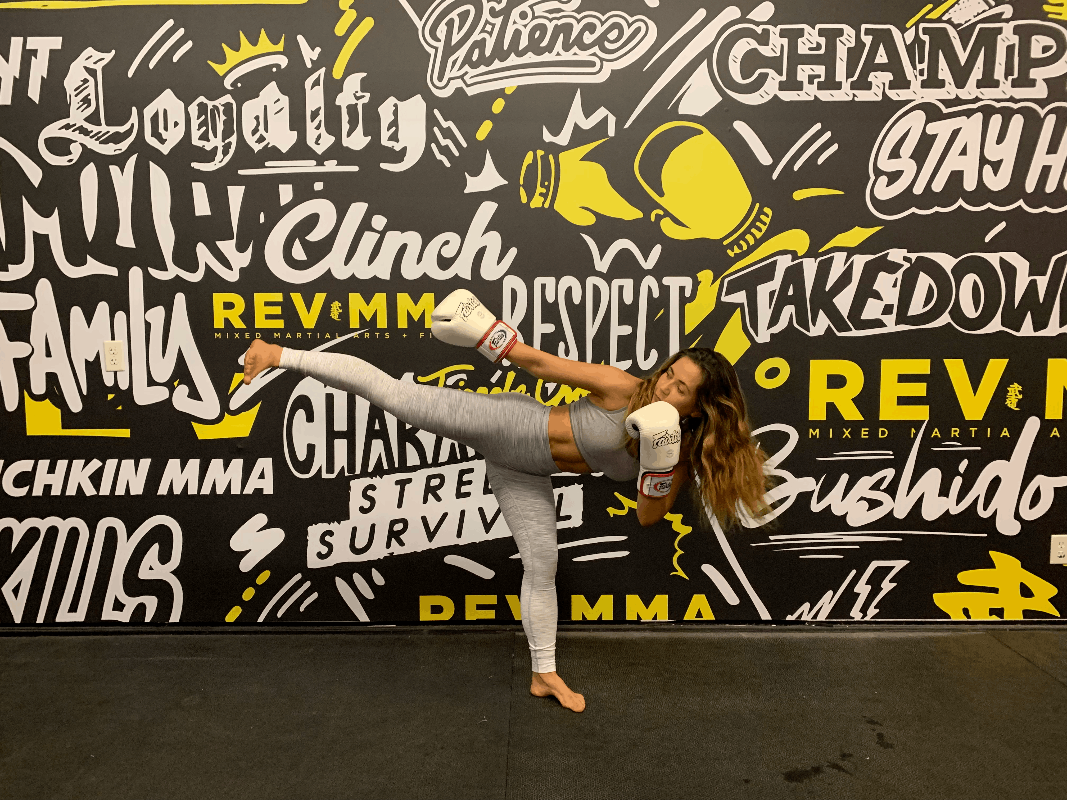 muay-thai-classes-north-york-revmma Female student throwing a high kick in boxing gloves in front of the REVMMA gym mural in North York Toronto