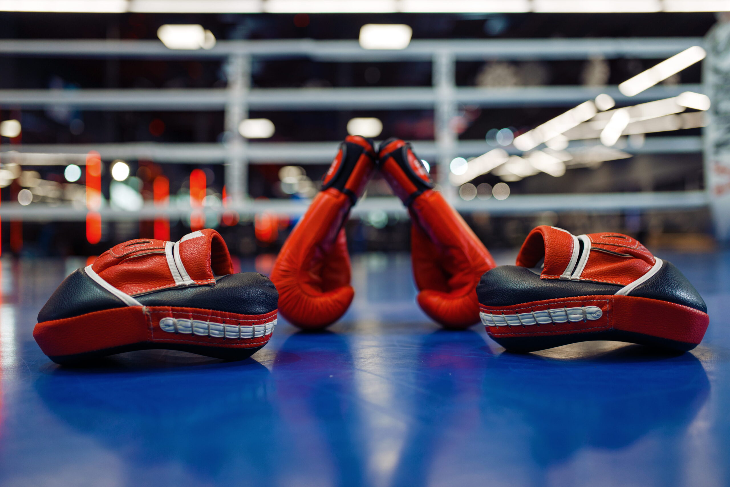 Red boxing gloves and focus pads on a blue boxing ring floor.