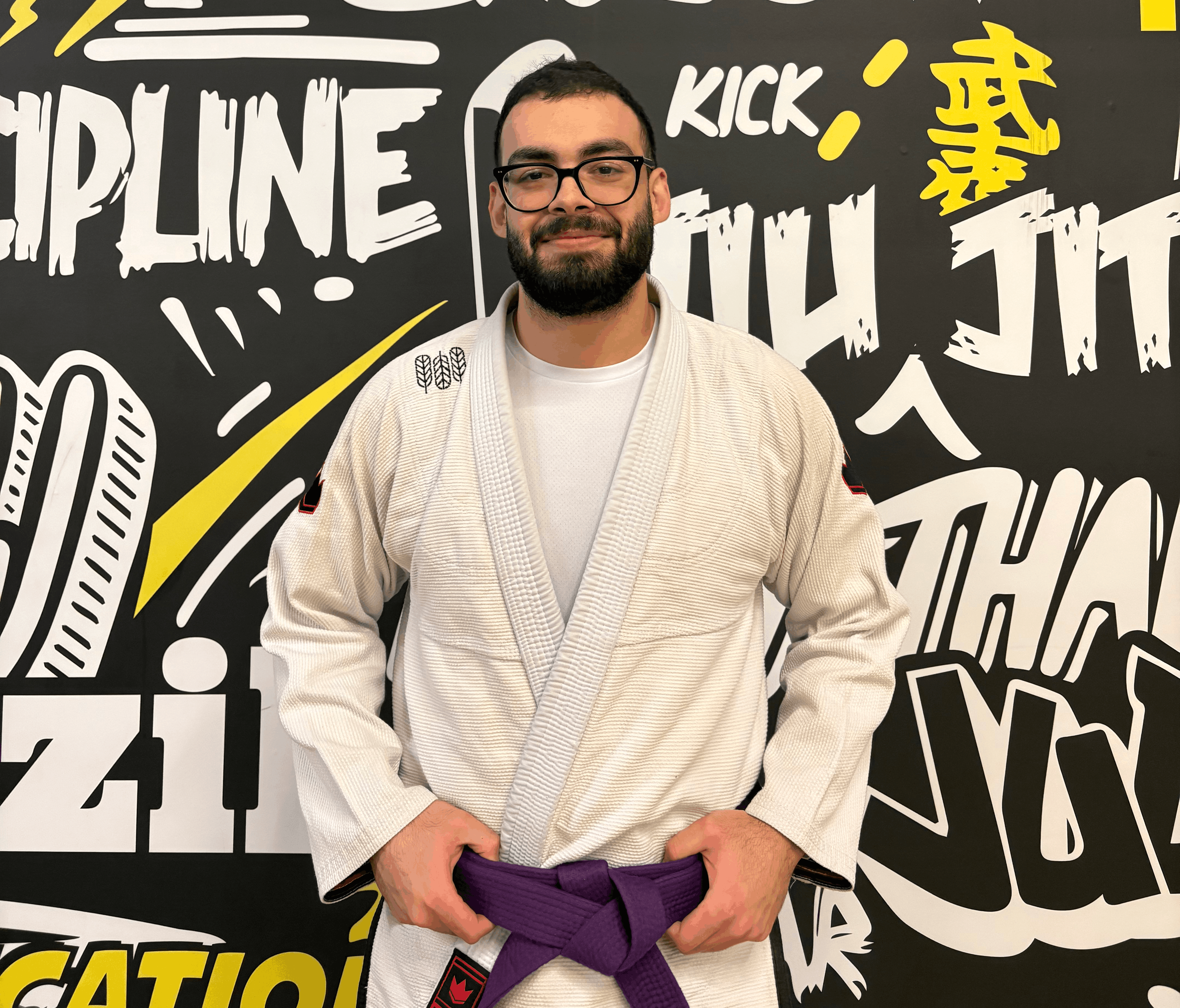Man in a white gi with a purple belt standing against a martial arts-themed graffiti wall.
