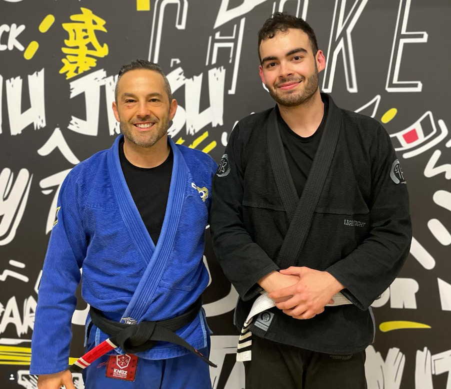 Two men in Jiu-Jitsu uniforms smiling in front of a graffiti wall.