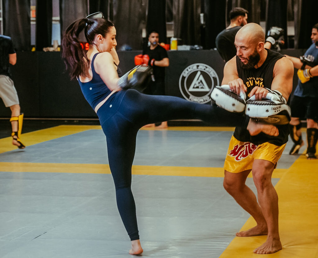 toronto-muay-thai-classes-revmma Female student practising a Muay Thai kick on focus mitts with an instructor on the mats at REVMMA Toronto
