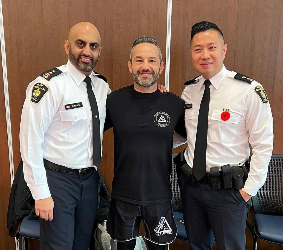 trusted-bjj-instructor-revmma Three men posing together, two in police uniforms, one in a black Gracie Jiu-Jitsu shirt, against a wooden background.